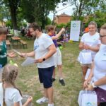 20250629_154045 Na zdjęciu grupa osób na zewnątrz wymienia się dyplomami. Tło to drzewa i budynki. Ubrania są jasne, dominują białe i zielone kolory.