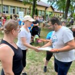 20250629_154126 Na zdjęciu jest grupa ludzi na zewnątrz, jedna osoba wręcza certyfikat innej. Tło to budynek i drzewa, kolorystyka jest naturalna. Ludzie są ubrani w letnie ubrania.