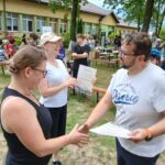 20250629_154128 Na zdjęciu są dwie osoby podające sobie ręce na tle budynku. W tle są ludzie siedzący na ławkach. Zieleń dominuje w krajobrazie wokół.