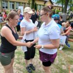 20250629_154142 Grupa ludzi na zewnątrz, kobiety podają sobie ręce. Zielona trawa, stoliki piknikowe w tle. Relaksująca, przyjacielska atmosfera.