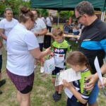 20250629_154222 Grupa osób w ogrodzie; dzieci w sportowych strojach z dyplomami, dorosły wręcza nagrody; zielony namiot w tle.