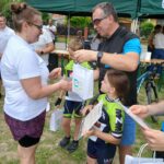 20250629_154226 Grupa ludzi rozdaje nagrody dzieciom w kolorowych strojach kolarskich. Tło to zielony namiot i trawnik. Przyjęcie odbywa się na świeżym powietrzu.