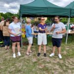 20250629_154416 Na zdjęciu widać grupę osób pod zielonym namiotem na świeżym powietrzu. Jedna osoba trzyma mikrofon, inna trzyma dyplom. Tło to zielone krzewy i niebo z chmurami.