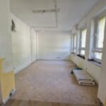 Pomieszczenie w trakcie remontu z jasnymi ścianami i beżową podłogą z płytek. Na prawej ścianie rząd dużych okien. Pod oknami podstawki pod kaloryfery i narzędzia.