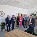 514538721_750997177275806_6176809876962503080_n Grupa ludzi stoi w jasnym pomieszczeniu biurowym. Na ścianie zegar i rośliny w tle. Osoby są ubrane elegancko.