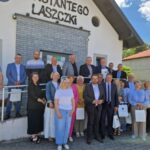 515058449_750997147275809_8681053122040016074_n Grupa ludzi na tle białego budynku z napisem. Jasne niebo, ceglana elewacja i torebki w dłoniach osób.