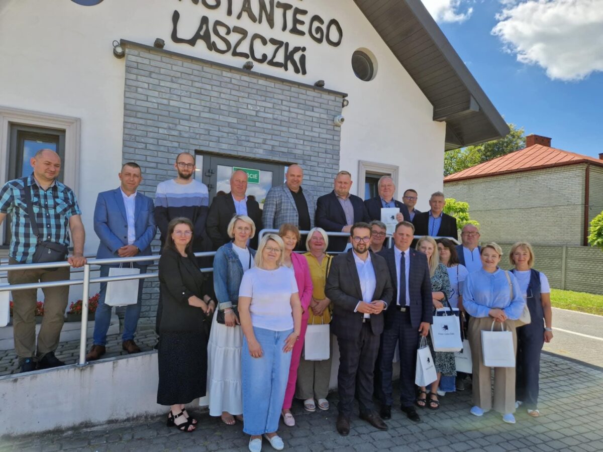 Grupa ludzi na tle białego budynku z napisem. Jasne niebo, ceglana elewacja i torebki w dłoniach osób.
