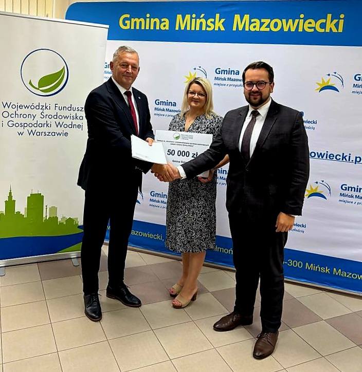Na zdjęciu troje osób w formalnych strojach trzymających duży czek. Tło to baner Gminy Mińsk Mazowiecki w bieli i niebieskim. Po lewej widoczny jest roll-up z logo ekologicznej organizacji.