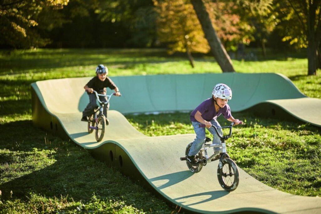 Dwóch dzieci na rowerach BMX jeździ po falistym torze na świeżym powietrzu. Tło stanowi zielona trawa i drzewa. Słońce rzuca cienie na tor.