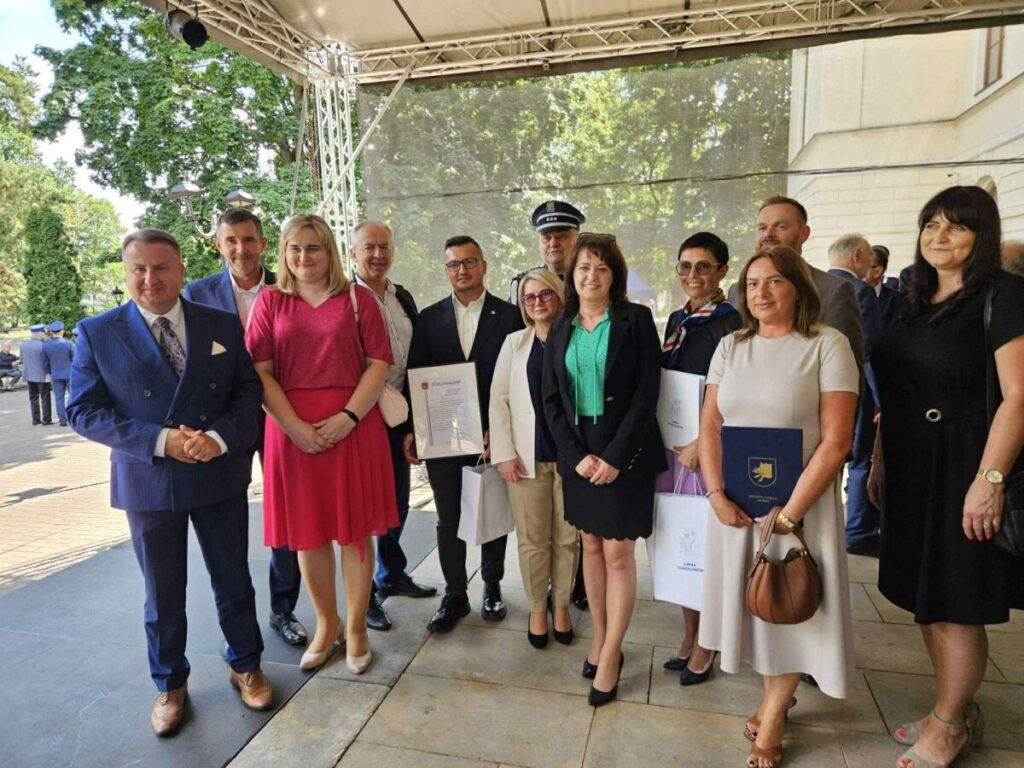 Na zdjęciu widoczni są ludzie stojący w grupie, ubrani elegancko. W tle zielone drzewa i białe ściany budynku. Niektóre osoby trzymają dokumenty i torby.