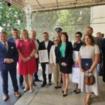 518375712_763804859328371_6214301076811974342_n Na zdjęciu widoczni są ludzie stojący w grupie, ubrani elegancko. W tle zielone drzewa i białe ściany budynku. Niektóre osoby trzymają dokumenty i torby.