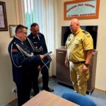 Trzech strażaków w mundurach stoi w biurze. Dwóch w ciemnych strojach ogląda dokument, jeden w jasnym stoi z boku. Za nimi jasne ściany i okno.