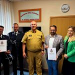 Grupa pięciu osób pozuje do zdjęcia w biurze. Dwie osoby w mundurach trzymają dokumenty, tło stanowi beżowa ściana z zegarem. Kolory: czerń, zieleń, beż.