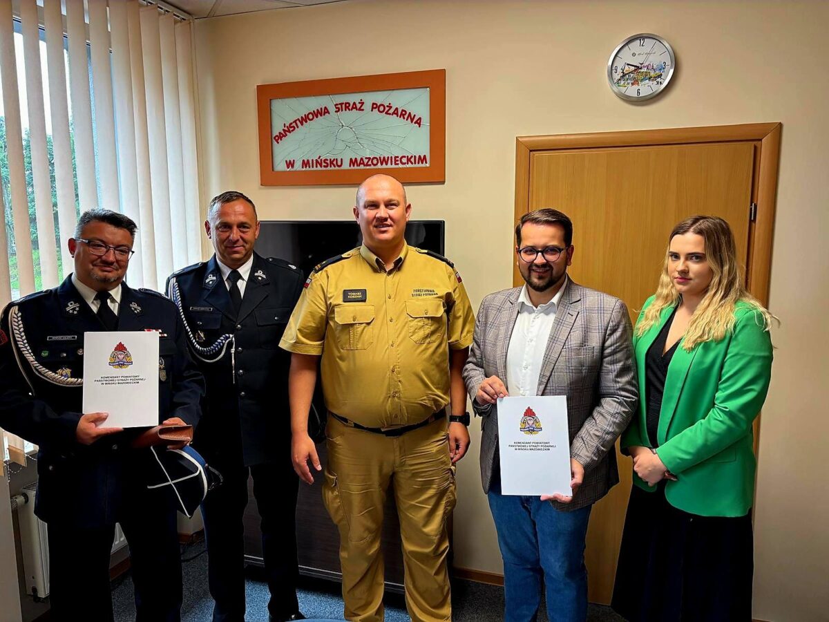 Grupa pięciu osób pozuje do zdjęcia w biurze. Dwie osoby w mundurach trzymają dokumenty, tło stanowi beżowa ściana z zegarem. Kolory: czerń, zieleń, beż.