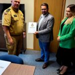 Trzy osoby stoją w biurze. Mężczyzna w mundurze, drugi w marynarce trzyma dokument, kobieta w zielonej marynarce obok. Tło to drzwi i szafka.