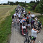 DSC_0001 Rowerzyści na ścieżce wśród pól, niektórzy prowadzą rowery, tło zielonych drzew i niebo z chmurami.