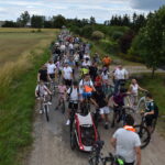 DSC_0002 Grupa ludzi na rowerach jedzie po wiejskiej drodze. Jasne niebo z chmurami, zielone pola po bokach. Tłum na drodze odświętnie ubrany.