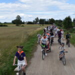 DSC_0003 Grupa rowerzystów jedzie polną drogą wśród pól pod niebieskim niebem. W tle widoczne są drzewa i budynki. Uczestnicy mają kolorowe kamizelki i różnorodne stroje.