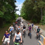DSC_0013 Grupa ludzi jedzie na rowerach wzdłuż asfaltowej drogi otoczonej zielenią drzew. Jest słoneczny dzień, w tle widać więcej uczestników. Po prawej stronie znajduje się chodnik i czerwona hydrantowa skrzynka.