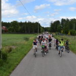 DSC_0014 Grupa rowerzystów jedzie wzdłuż wiejskiej drogi. Otacza ich zielone pola i drzewa. Niebo jest częściowo pochmurne.