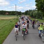 DSC_0015 Grupa rowerzystów jedzie drogą polną, otoczoną zielonymi polami. W tle las i jasne niebo z chmurami. Przeważające kolory to zieleń, błękit i szarość.