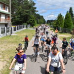 DSC_0016 Ulicą w lesie jedzie grupa rowerzystów w kolorowych ubraniach. W tle dom i zielone drzewa. Niebo jest częściowo zachmurzone.