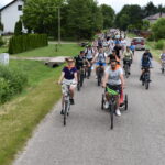 DSC_0019 Na zdjęciu widoczna jest grupa rowerzystów na wiejskiej drodze. Otoczenie to zielone pola i drzewa, w tle domy. Ludzie mają kolorowe stroje, a pogoda jest słoneczna.