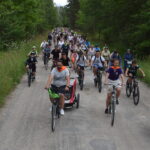 DSC_0023 Grupa rowerzystów na leśnej ścieżce, otoczonej zielenią. Uczestnicy w kolorowych kaskach i strojach. Pogoda zachmurzona, tło z drzewami.