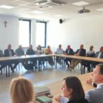 IMG_20250630_110140 Zdjęcie przedstawia grupę ludzi siedzących przy dużym prostokątnym stole w jasnym pomieszczeniu. Na ścianie w tle widać krzyż i okna z jasnym światłem. Osoby noszą formalne ubrania, a oczy są zwrócone w jednym kierunku.