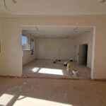 Pomieszczenie w remoncie z jasnobeżowymi ścianami i otwartymi oknami. Na podłodze są puszki z farbą. W tle drzwi i puste ściany.