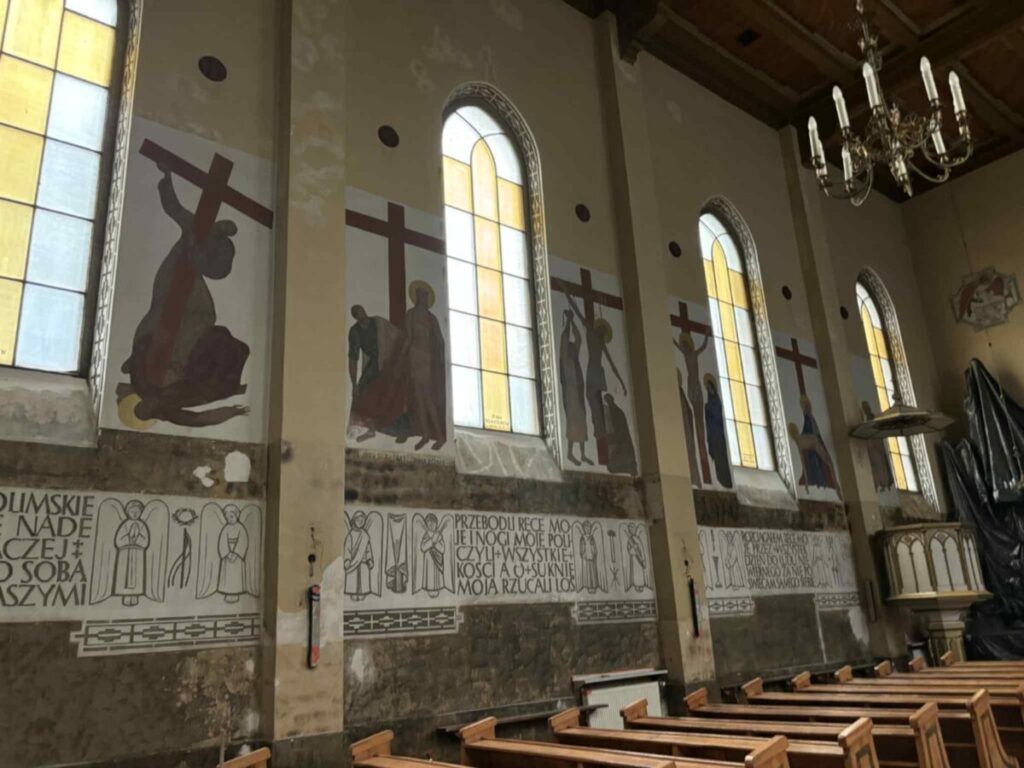 Wnętrze kościoła z drewnianymi ławkami. Na ścianie wiszą malowidła stacji Drogi Krzyżowej pod dużymi, żółtymi oknami. Dekoracyjne napisy u dołu ściany i żyrandol na drewnianym sufic