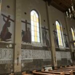 Wnętrze kościoła z drewnianymi ławkami. Na ścianie wiszą malowidła stacji Drogi Krzyżowej pod dużymi, żółtymi oknami. Dekoracyjne napisy u dołu ściany i żyrandol na drewnianym sufic