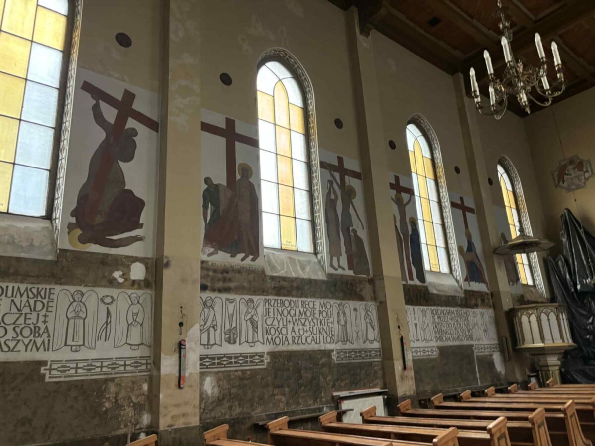 Wnętrze kościoła z drewnianymi ławkami. Na ścianie wiszą malowidła stacji Drogi Krzyżowej pod dużymi, żółtymi oknami. Dekoracyjne napisy u dołu ściany i żyrandol na drewnianym sufic