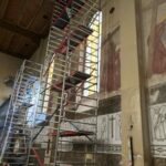 Wnętrze kościoła z freskami na ścianach w odcieniach brązu i szarości. Na pierwszym planie stoją metalowe rusztowania. Na suficie widoczne drewniane belki.