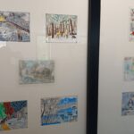 Na obrazie widoczne są kolorowe rysunki w ramkach na białym tle. Przedstawiają zimowe krajobrazy z domkami i lasami. Całość jest oszklona i odbicie okien można dostrzec na powierzchni.