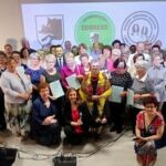 Grupa osób różnej wiekowo, uśmiechnięta, pozująca do zdjęcia w sali. W tle widoczne logo z połączeniem kolorów zieleni oraz bieli. Większość trzyma dokumenty.