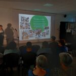 Sala prezentacyjna z ciemnym tłem, projektorem i kobietą przy mównicy. Ekran wyświetla kolorowe ilustracje. Publiczność siedzi na krzesłach.