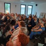 Grupa osób siedzi w pomieszczeniu z jasnymi ścianami, na białych krzesłach. Na ścianach wiszą obrazy i znajduje się krzyż. Okna wpuszczają naturalne światło.