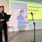 Na zdjęciu mężczyzna w garniturze przemawia do mikrofonu. Obok niego widoczna jest jasna ściana z zielono-żółtym ekranem z napisem Uniwersytet Dobrego Wieku. Pomieszczenie jest dobrze oświetlone, a tło jest neutralne.