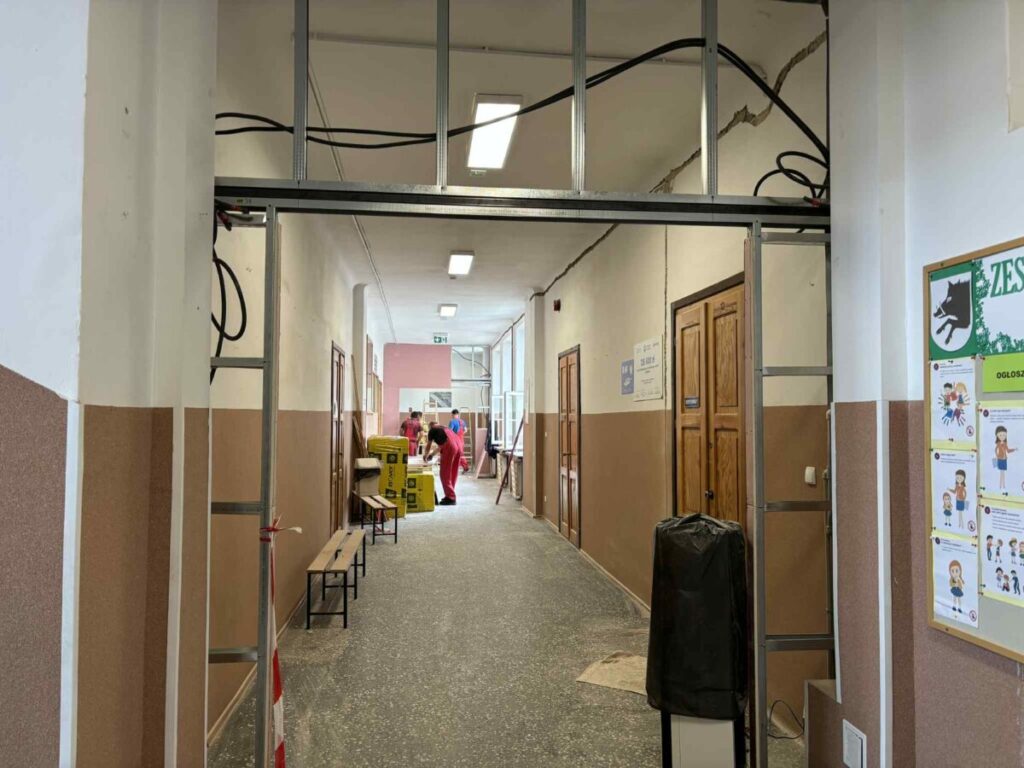 Korytarz szkolny w trakcie remontu, z pracownikami i narzędziami. Ściany biało-brązowe, podłoga szara. Widoczne oznaki napraw.
