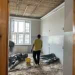 Pomieszczenie w remoncie z drewnianym sufitem i szarymi ścianami. Mężczyzna w żółtej koszulce i błękitne wiadro na podłodze. Duże okno wpuszcza światło dzienne.