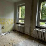 Pomieszczenie w trakcie remontu z żółtymi ścianami i gruzem na podłodze. Dwa duże okna z widokiem na drzewa. Biały grzejnik pod oknami.