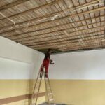 Na zdjęciu osoba stoi na drabinie, malując sufit z cegieł. Ściany są białe i żółte, wnętrze wygląda na remontowane. Tło jest proste, bez mebli.