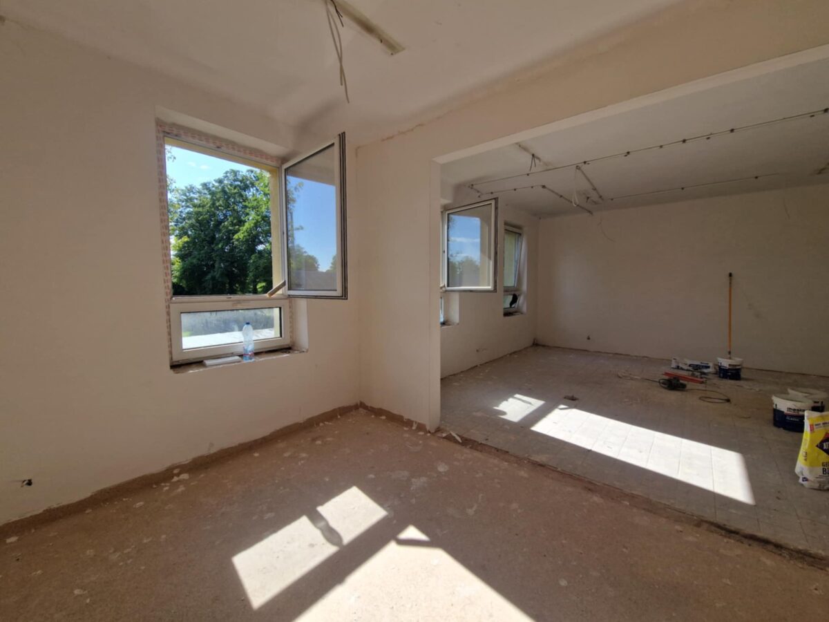 Pomieszczenie w remoncie o białych ścianach, z otwartymi oknami. Podłoga z plamami farby, materiałami i sprzętem. W tle widać zielone drzewa.