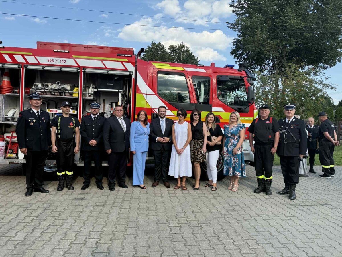 Grupa ludzi stoi przed czerwonym wozem strażackim na tle zielonych drzew i błękitnego nieba. Widać strażaków i osoby w eleganckich ubraniach. Tło jest jasne i słoneczne.