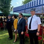 Grupa osób stoi na trawie pod niebiesko-białym namiotem. Mężczyźni i kobieta ubrani są formalnie, z tyłu widać drzewa i krzesła. Jedna osoba ma na sobie mundur.