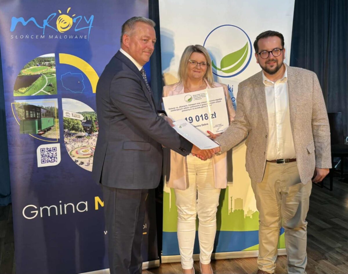 Trzech ludzi na tle plakatów z logo Mrozów, ściska dłonie. Osoba z certyfikatem w ręku. Dominują kolory niebieski i zielony.