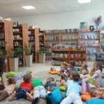 Grupa dzieci leży na poduszkach w bibliotece, słuchając dorosłej osoby. Tło stanowią drewniane regały pełne książek. Kolorowe poduszki i zabawki tworzą przytulną atmosferę.