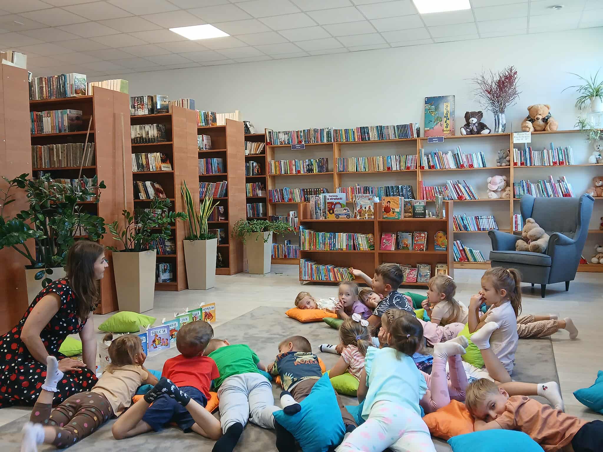 Grupa dzieci leży na poduszkach w bibliotece, słuchając dorosłej osoby. Tło stanowią drewniane regały pełne książek. Kolorowe poduszki i zabawki tworzą przytulną atmosferę.