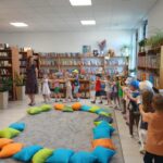 Grupa dzieci bawi się w sali z drewnianymi półkami pełnymi książek. Na podłodze leżą kolorowe poduszki. Ściany są jasne, a wnętrze jest dobrze oświetlone.
